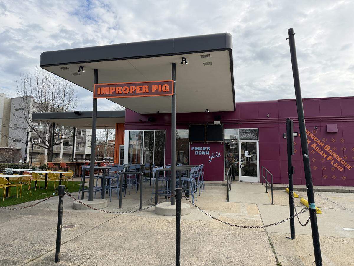 An exterior shot of The Improper Pig restaurant featuring a purple facade, an orange “Improper Pig” sign under a large overhang, and an outdoor patio with blue and yellow seating.