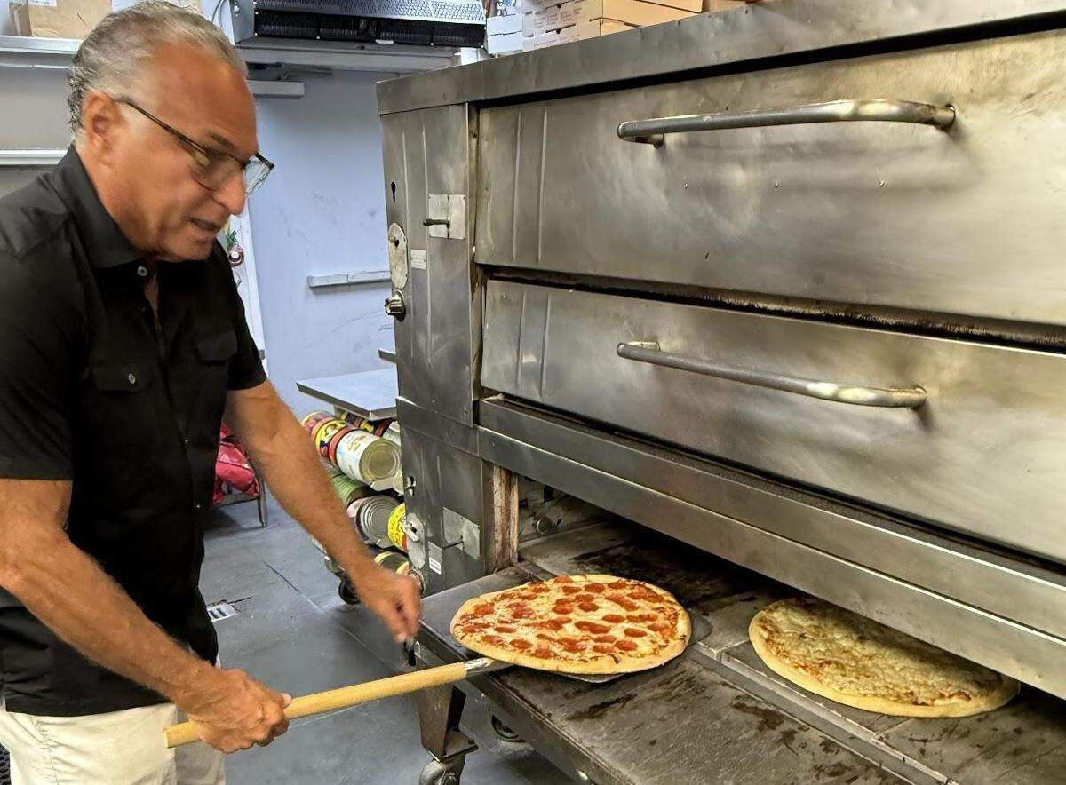 A male chef with glasses and a black collared shirt is using a long wooden pizza peel to remove a freshly baked pepperoni pizza from a large, industrial-style pizza oven. Another cheese pizza is visible next to it, still in the oven. The oven is a large, stainless steel appliance with two sections, and the background shows a storage area with cans and other restaurant supplies.
