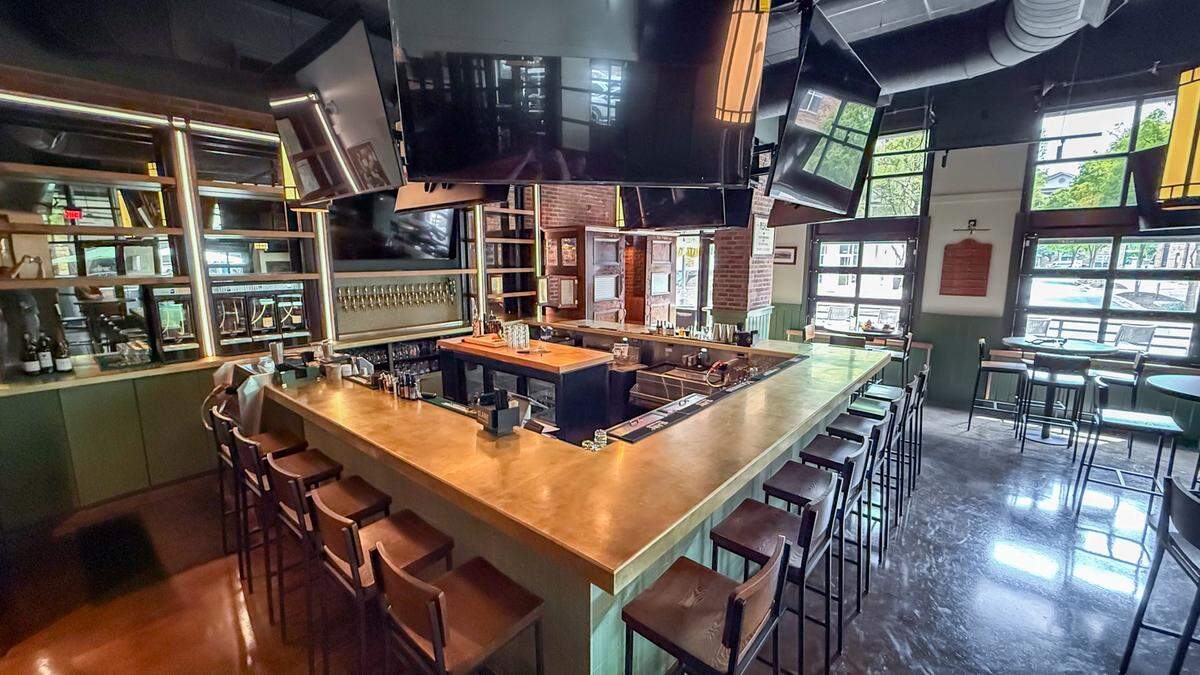 A wide-angle view of the primary bar area. The bar features a burnished gold L-shaped countertop lined with dark wood barstools. Behind the bar, a large bank of beer taps is visible against a brick wall, and several large flat-screen TVs are suspended from the dark, industrial ceiling. Large windows in the background let in plenty of natural light.