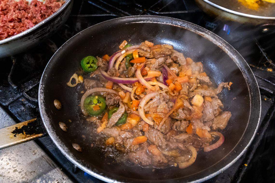 A close-up, high-action shot of a sauté pan on a commercial burner. Inside the pan, strips of beef Tibs are being seared with sliced red onions, tomatoes, and fresh green jalapeños. Steam rises from the pan, and the glistening oil suggests a flavorful, savory dish in progress.
