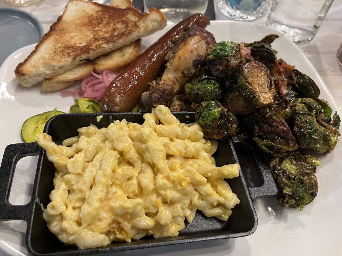 A high-angle, close-up shot captures a white plate piled with a barbecue-style meal. In the foreground, a small, black rectangular dish overflows with creamy macaroni and cheese. To the right is a large serving of dark, roasted Brussels sprouts, and in the background are a smoked sausage link, a piece of roasted chicken, and thick-cut toast.