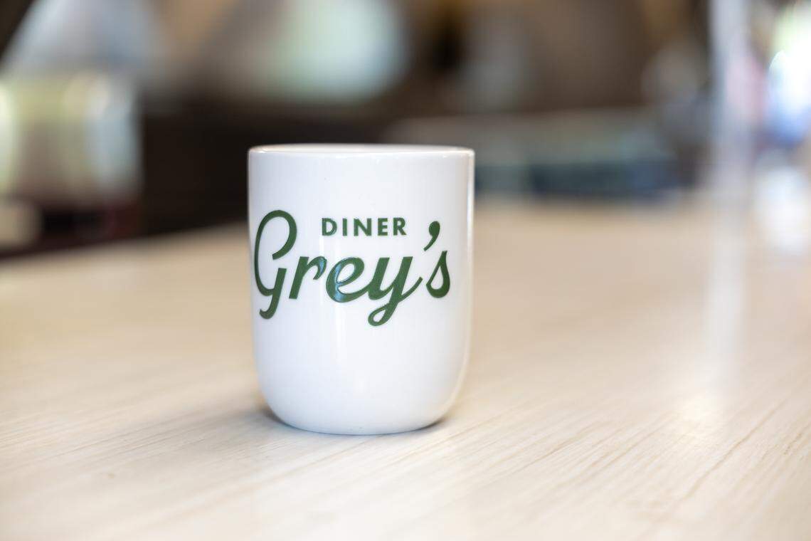 A close-up, shallow-focus shot of a white ceramic mug sitting on a light-colored wooden counter. The mug features the “Grey’s DINER” logo in a forest green script and block font. The background is softly blurred, suggesting the interior of a diner.