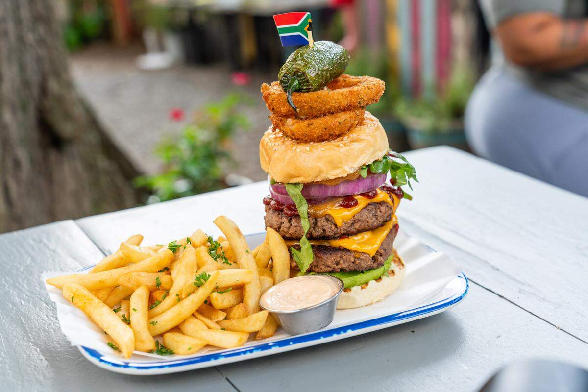 A massive vegan double cheeseburger and a side of golden French fries sit on a white plate on a patio table. The towering burger is topped with bright red onion, melted cheese, two crispy onion rings, and a whole fried jalapeño, all held together by a toothpick with a small South African flag.