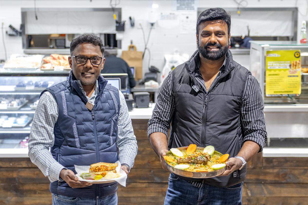 A medium, eye-level shot of two restaurant owners standing side-by-side and smiling, appearing to be in a food market or eatery. The one on the left, wearing glasses and a blue vest, holds a paper plate with a sandwich or pastry. The one on the right, wearing a black vest, holds a large, round metal platter piled with rice, meat, and vegetables. Behind them, refrigerated food display cases and a service counter are visible.