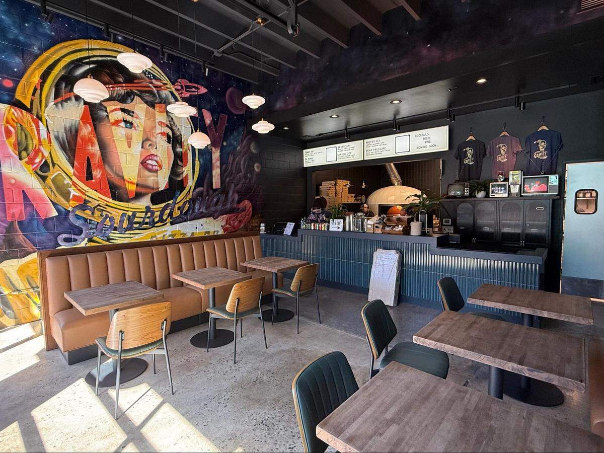 A wide shot of the interior of a modern, space-themed restaurant, likely Gravity Sourdough Pizza. On the left, a massive, colorful mural of a retro, pop-art-style female astronaut in a helmet dominates the wall above a long, tan-colored booth. The restaurant features wooden tables with black bases, paired with modern chairs. In the background, a service counter with a ribbed, dark-teal base and a light-colored top sits in front of the kitchen area, where the large, cream-colored dome of a pizza oven is visible. A menu board is mounted on the wall above the counter.