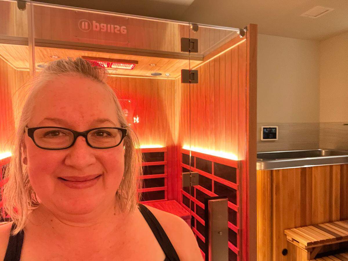 A high-angle, medium-close-up selfie of a person with light skin, grey hair and black-rimmed glasses, smiling at the camera and wearing a black tank top. To the left is an infrared sauna with glass doors and wood-paneled interior walls. To the right, behind a glass partition, there is a cold plunge tub made of matching wood with a metal rim.