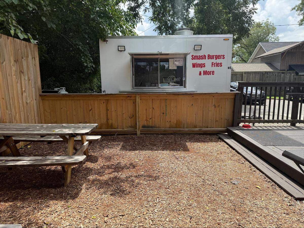 A small, white food trailer with the words “SMASH BURGERS, Wings, Fries & More” written in red is set up behind a light wooden fence. In front of the trailer, there is an outdoor seating area with wood chips on the ground and a wooden picnic table. A wooden deck and fence are visible on the right, and trees and a house are in the background.