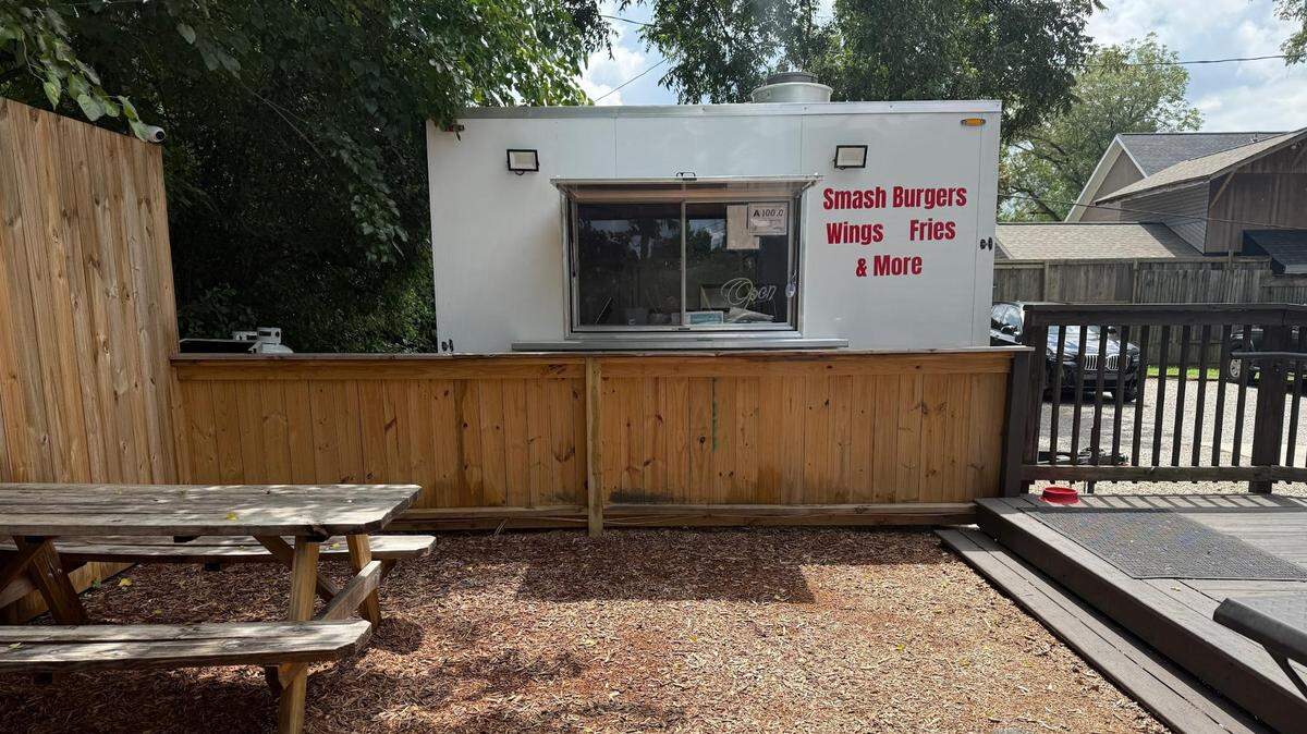 A small, white food trailer with the words “SMASH BURGERS, Wings, Fries & More” written in red is set up behind a light wooden fence. In front of the trailer, there is an outdoor seating area with wood chips on the ground and a wooden picnic table. A wooden deck and fence are visible on the right, and trees and a house are in the background.