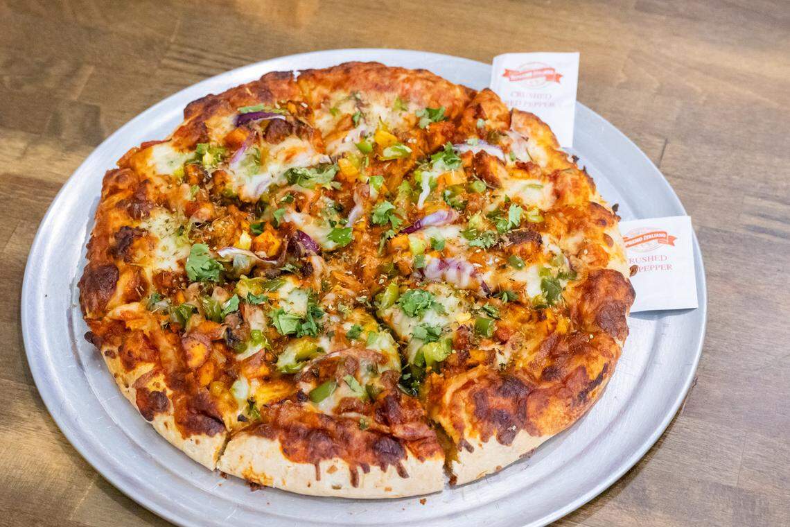 A close-up view of a whole, uncut pizza on a metal serving tray. The pizza has a thick, browned crust and is topped with mozzarella cheese, red onion, green peppers, and generous amounts of cilantro over a red sauce base. Two small packets of crushed red pepper flakes are visible on the side.