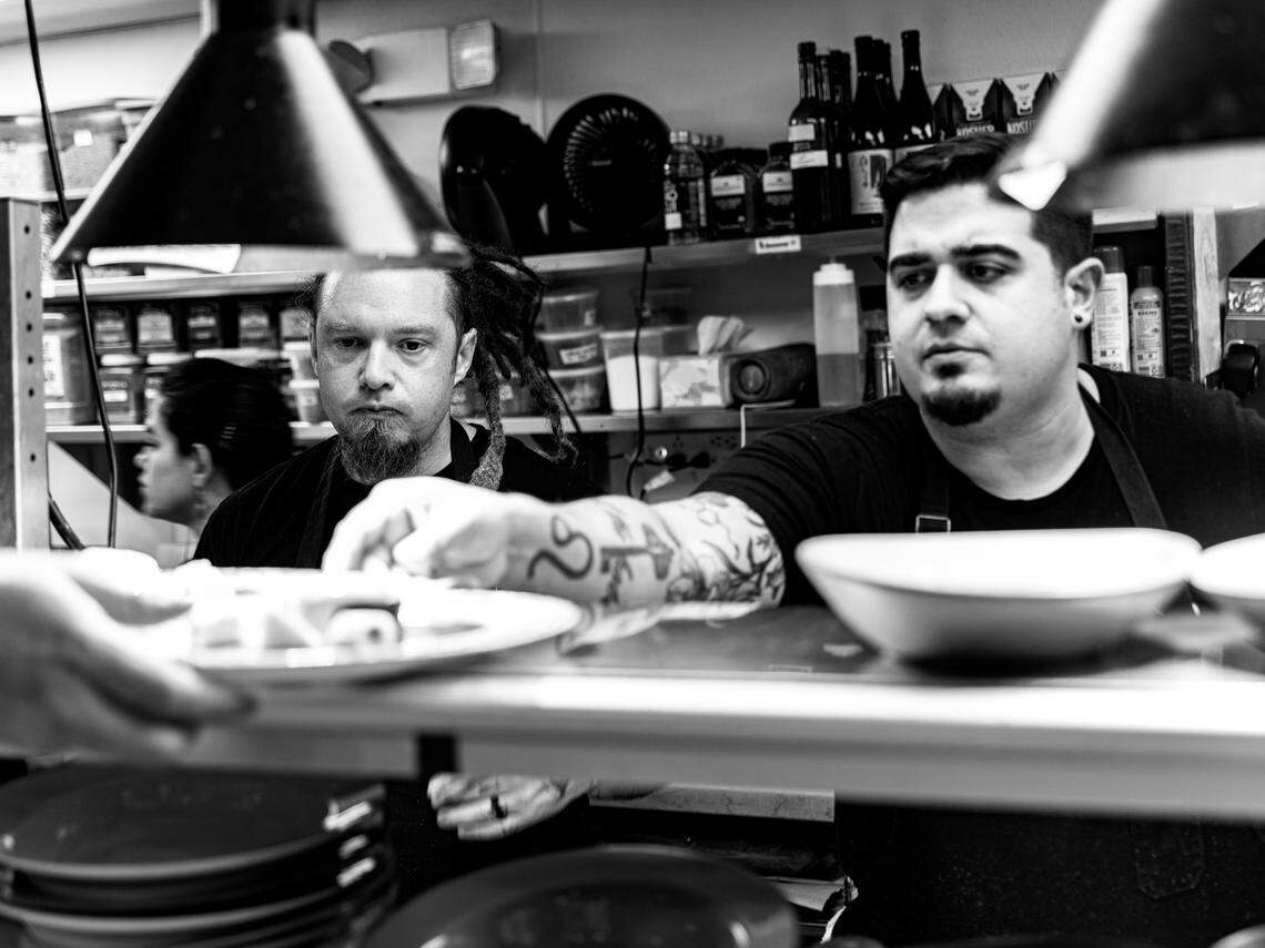 In a dynamic black and white shot taken from the perspective of the service counter, two chefs work at a kitchen pass. One chef with a tattooed forearm arranges food on a plate, his expression focused. A second chef with dreadlocks and a goatee looks on intensely from behind. The foreground and background show the bustling environment of a professional kitchen.