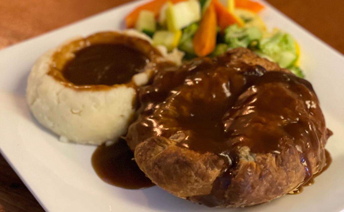 A close-up shot of a plated meal on a white plate, featuring a golden-brown meat pie and a scoop of mashed potatoes, both generously covered in dark gravy. A side of colorful steamed vegetables, including broccoli, carrots, and squash, is visible in the background.