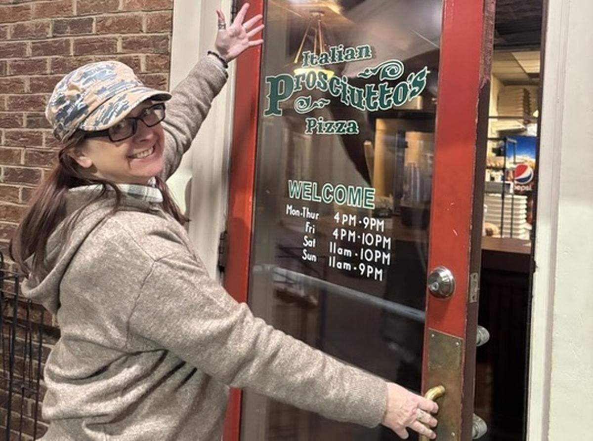 An image showing a person with long brown hair and wearing a camouflage baseball cap, glasses and a tan hoodie standing in front of the entrance to a restaurant called Prosciutto’s Italian Pizza. One hand is on the gold door handle and the other is raised in a wave.