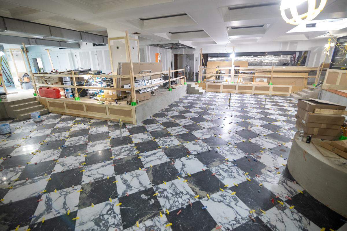 The interior of a large commercial space under renovation. The floor is covered in a checkered black-and-white marble tile with yellow and orange tile spacers still in place. Temporary wooden railings and construction supplies are scattered throughout the room.