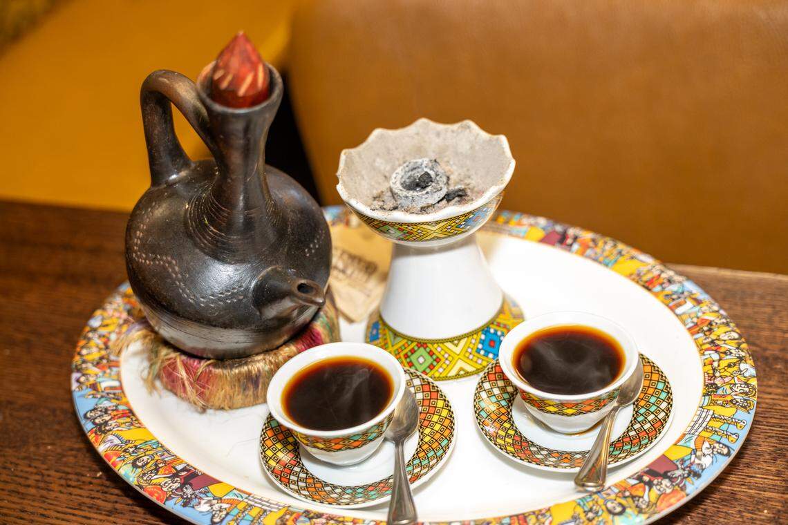 A close-up of a traditional Ethiopian coffee ceremony set on a colorful decorative tray. The set includes a Jebena (a handmade black clay coffee pot), two small ceramic cups filled with dark coffee, and a white incense burner releasing a thin trail of smoke from a glowing coal. The tray and cups are adorned with bright, geometric Ethiopian patterns, emphasizing the cultural significance of the coffee ritual.