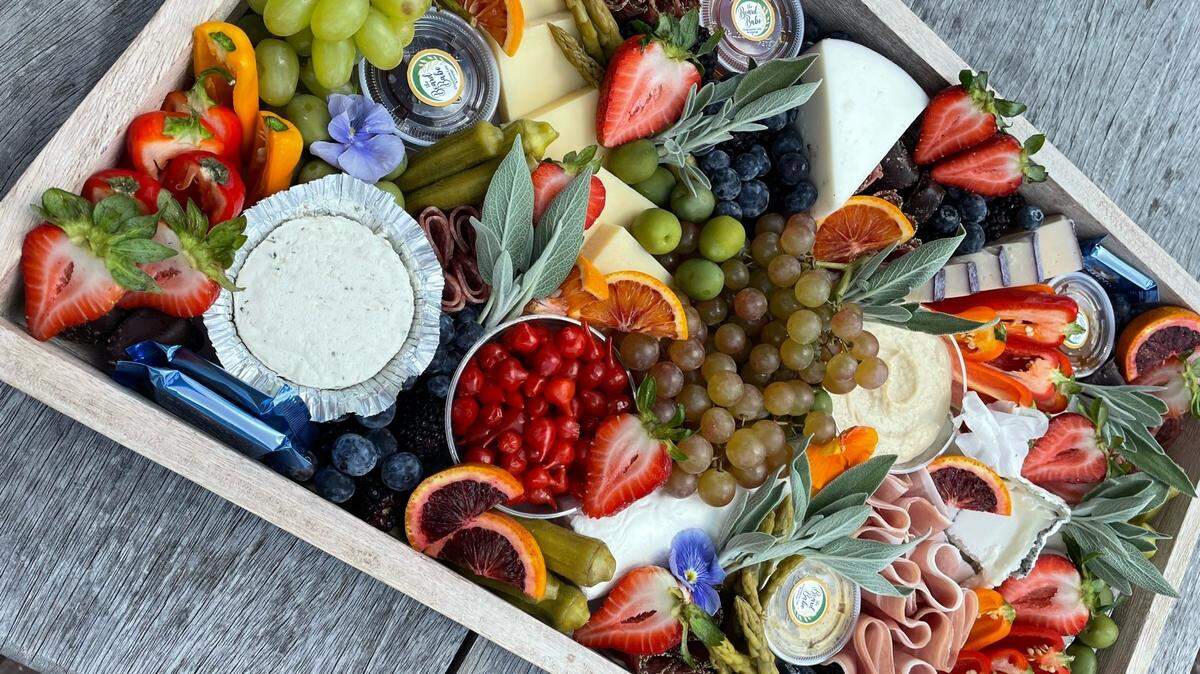 A close-up, overhead view of a wooden rectangular tray filled with an artistic and colorful arrangement of food. The tray is densely packed with a variety of fruits like grapes, strawberries, and oranges, along with different cheeses, sliced meats, and dips, set on a rustic wooden table.