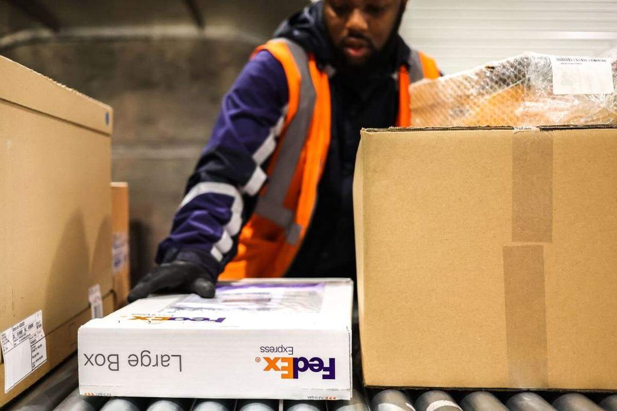 A package handler wearing a black jacket and an orange safety vest guides a large FedEx-branded box moving on a conveyor belt. The foreground is filled with several cardboard boxes, indicating a busy warehouse or package sorting facility environment.