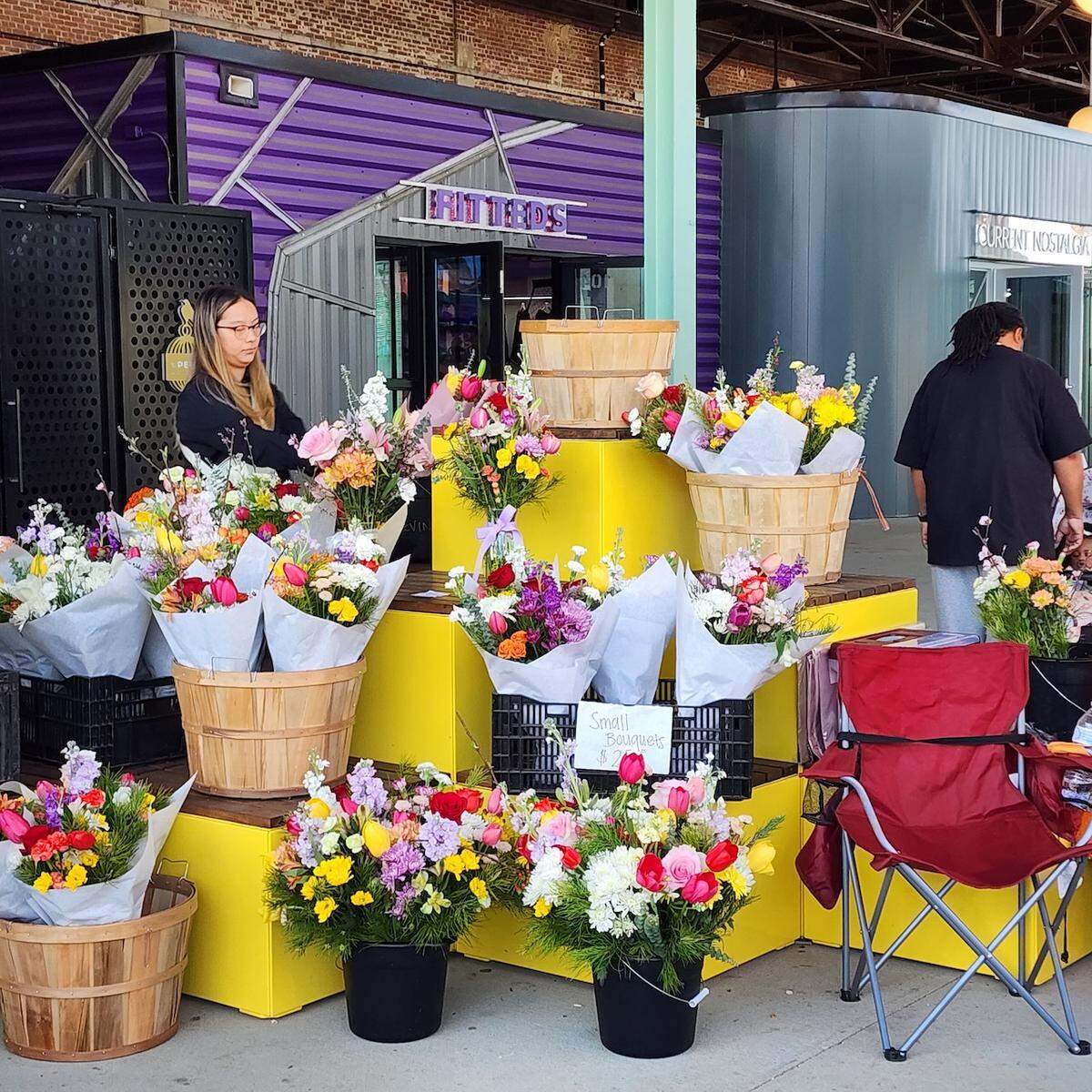 A vendor stands behind a bright yellow outdoor flower stall selling colorful bouquets. The flowers, including tulips and other mixed varieties, are presented in wooden bushel baskets and black buckets, wrapped in white paper. The stall is set up in a covered outdoor market area with purple and gray structures in the background.