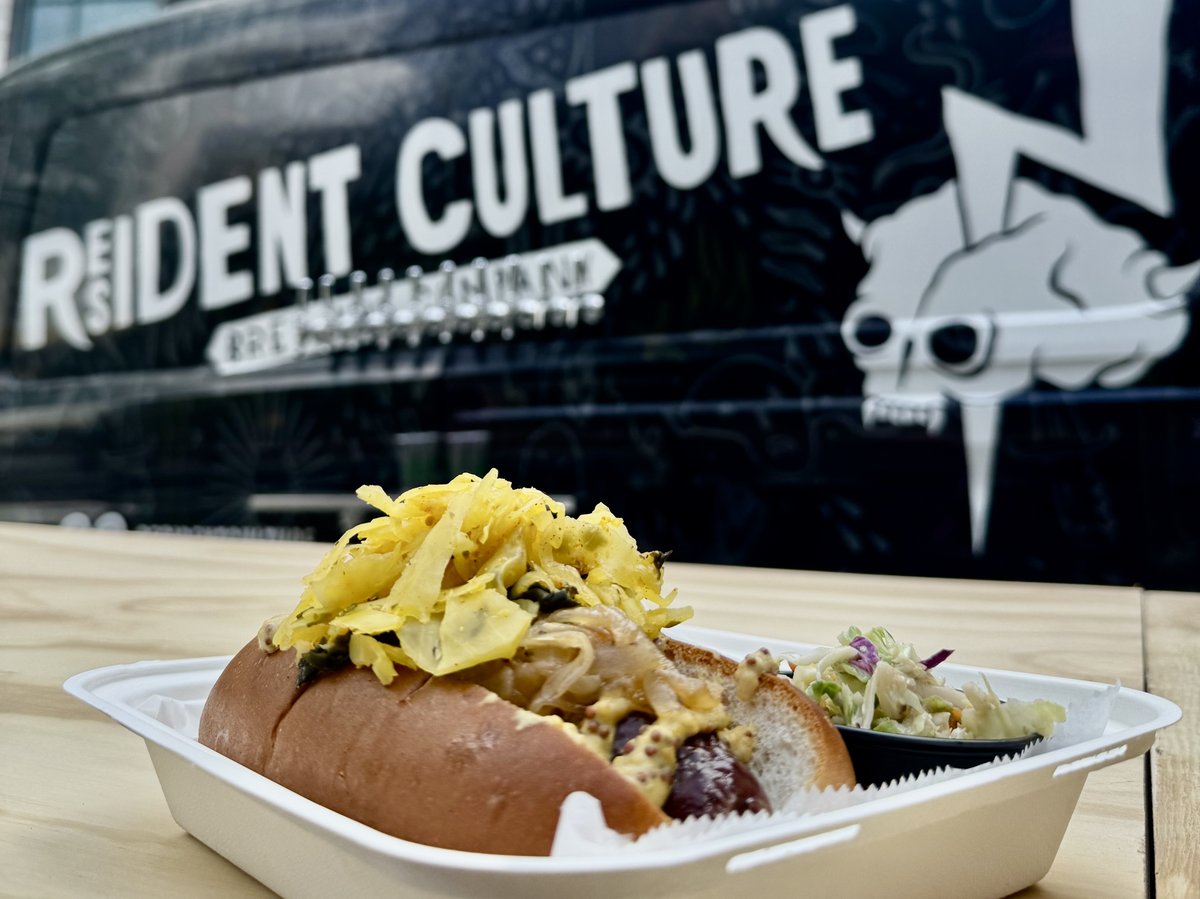 A low-angle photo centers on a loaded bratwurst in a takeout container with a side of coleslaw. In the soft-focus background, a black van is clearly branded with the white text “Resident Culture Brewing Company” and a graphic of a skull with a lightning bolt.