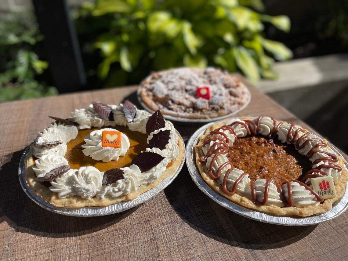 Three holiday pies sitting on a wooden table outdoors in the sun. The pie in the foreground left is a pumpkin or sweet potato pie decorated with whipped cream swirls, chocolate leaves, and a small square tag that says “thanks.” The pie on the right is a pecan pie with a decorative ring of whipped cream and caramel drizzle. The pie in the background is a fruit crumble pie, possibly apple, topped with powdered sugar and a small red tag. The background features blurred green foliage.