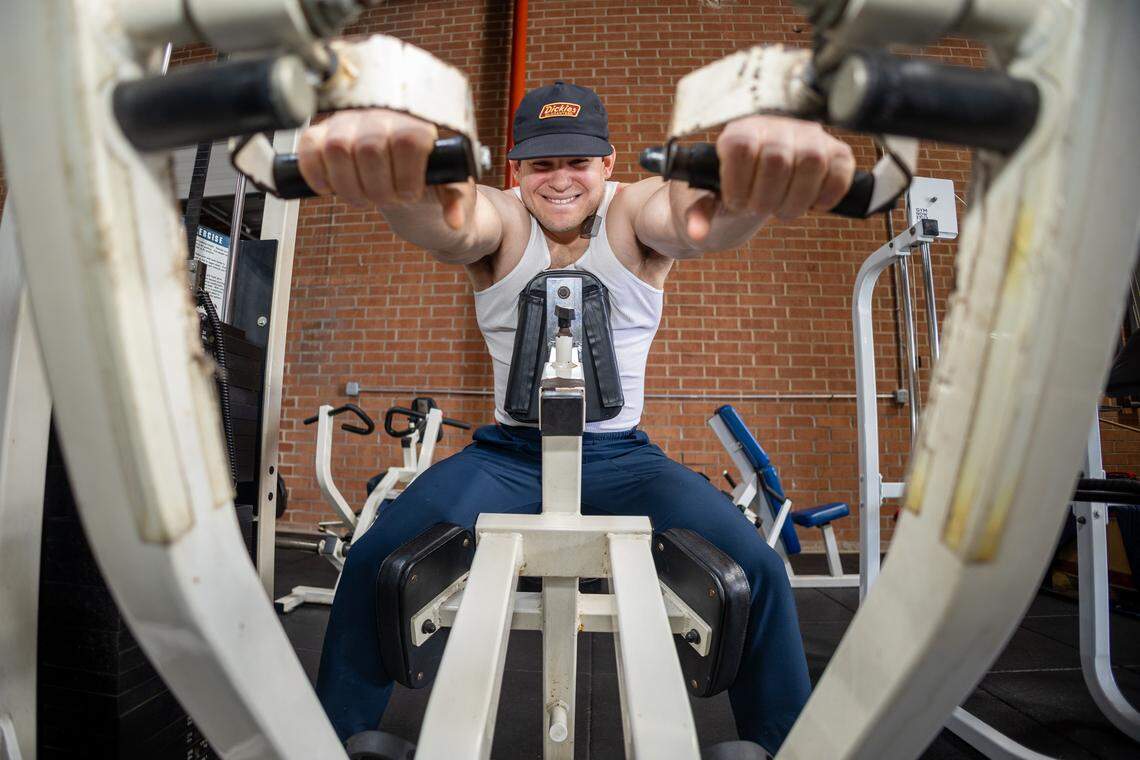 Gym user in a white tank top and a dark Dickies baseball cap performs a seated press on a white, vintage weight machine with a red brick wall background.
