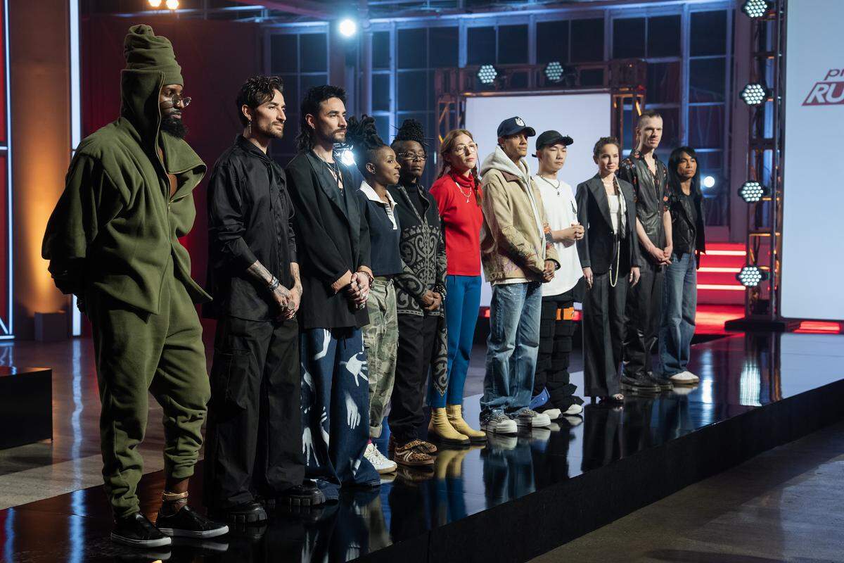 A lineup of eleven diverse fashion designers on a stage. They stand in a row on a brightly lit runway, looking off to the side as if at a panel of judges. The designers are wearing a variety of unique and personal styles, from streetwear to avant-garde. The “Project Runway” logo is partially visible on a large screen in the background.