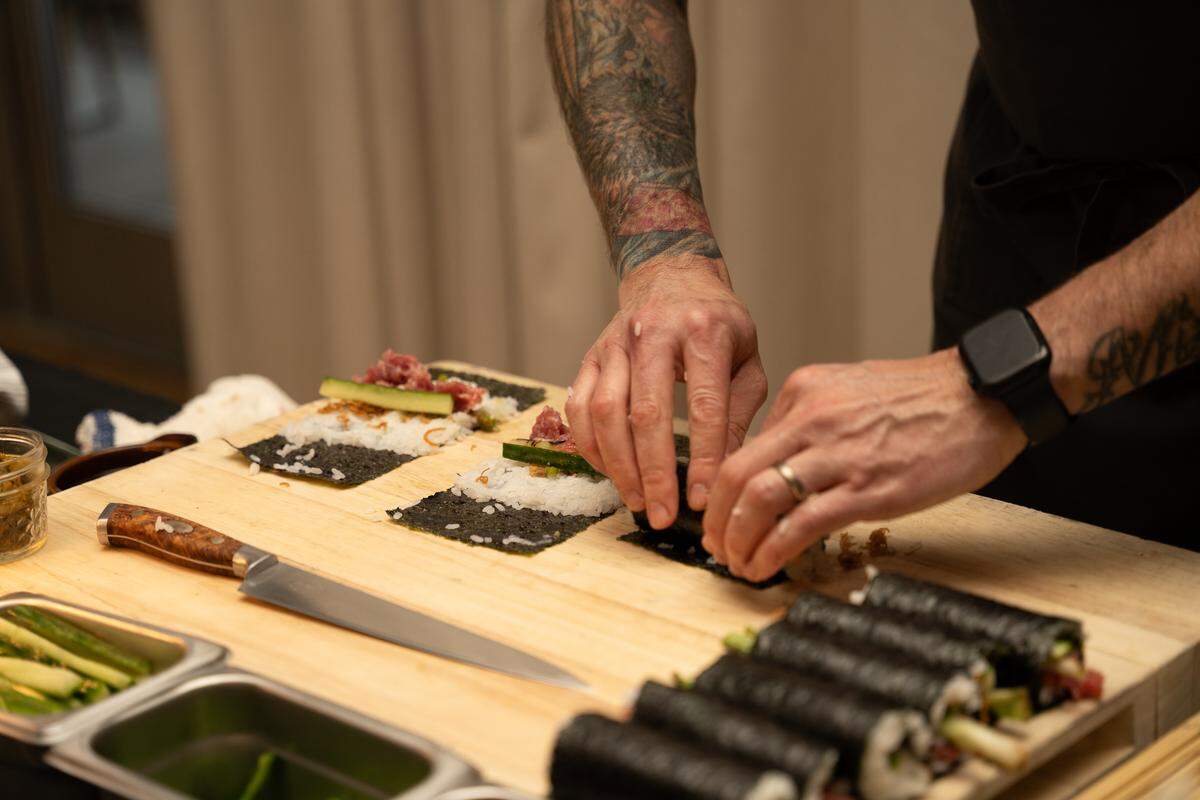 A chef’s tattooed hands are shown rolling sushi on sheets of nori and rice atop a wooden board. Several completed sushi rolls are lined up in the foreground.