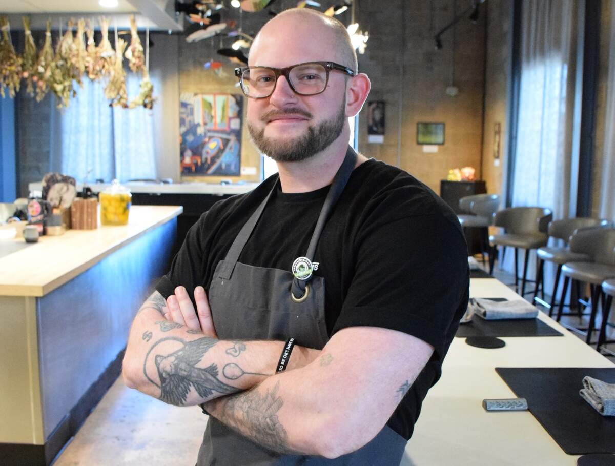 An eye-level portrait of a bald chef with glasses and a well-groomed beard, standing with folded arms inside a modern restaurant. The chef has a nose piercing and prominent tattoos on both forearms, and is wearing a black t-shirt under a dark gray chef’s apron featuring a small green pin on the strap. In the background, a sleek, light-colored bar counter with under-counter blue lighting is visible on the left, while a long dining counter lined with minimalist gray barstools stretches along the right. The warm interior is decorated with hanging dried herbs, artwork, and concrete-style walls.