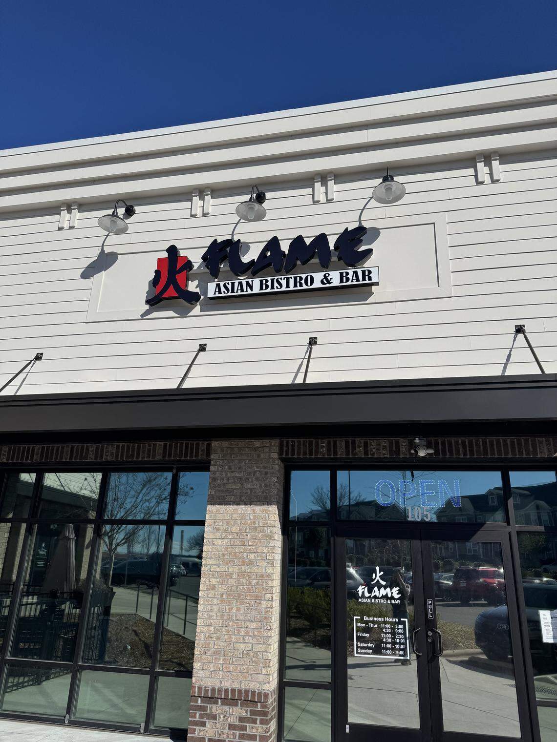 Exterior of Flame Asian Bistro & Bar. The building features white horizontal siding, a black awning, and a modern sign with a red “fire” kanji character. A brick pillar stands between large glass windows and the entrance door, which displays the restaurant’s business hours.