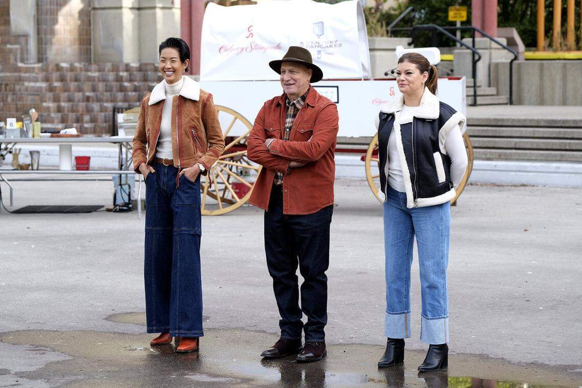 A full-length photo of the three Top Chef hosts outdoors. Kristen Kish wears a brown suede jacket, Tom Colicchio is in a rust-colored jacket and a hat, and Gail Simmons wears a black shearling-lined vest. They are smiling and standing in front of a large, white covered wagon, suggesting an on-location challenge.