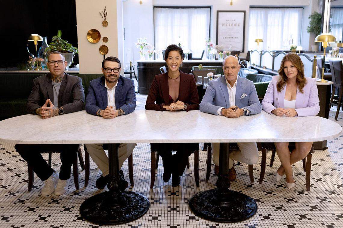 The image shows five judges seated behind a long, white marble-top table at a restaurant, including a person with hand and neck tattoos wearing glasses and a dark grey blazer, a person with a beard and glasses wearing a blue patterned blazer, a person wearing a dark maroon blazer, a bald person wearing a light blue blazer over a white shirt and a person with reddish-brown hair,wearing a light purple blazer. The setting is an upscale dining room with a black and white tiled floor, green velvet booth seating in the background, and various floral arrangements and gold lamps.