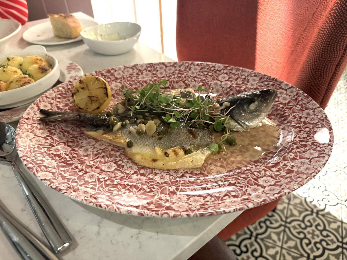 A whole roasted or grilled fish served in a shallow pool of light sauce on a red-and-white floral patterned oval platter. The fish is presented head-on, topped with capers, slivered garlic, and fresh green microgreens. A charred lemon half rests on the side of the plate.