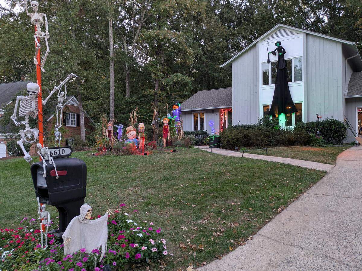 A wide shot of a suburban house’s front yard, extensively decorated for Halloween at dusk. The lawn is filled with a variety of spooky figures, including scarecrows and ghouls, some lit with red lights. On the right, a large witch figure is mounted on the house above a window, which glows with green light. In the foreground by the driveway, a tall orange pole next to the mailbox is covered in climbing skeletons, and a small ghost figure rises from a flower bed.
