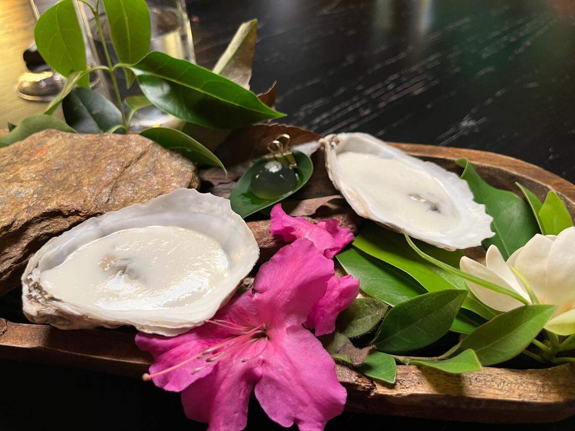 A deconstructed oyster dish from a tasting menu. Two shells holding a smooth, white, creamy substance are presented in a dark wood bowl. The presentation is highly naturalistic, with the shells arranged amongst a real rock, glossy green leaves, and fresh pink and white flowers.
