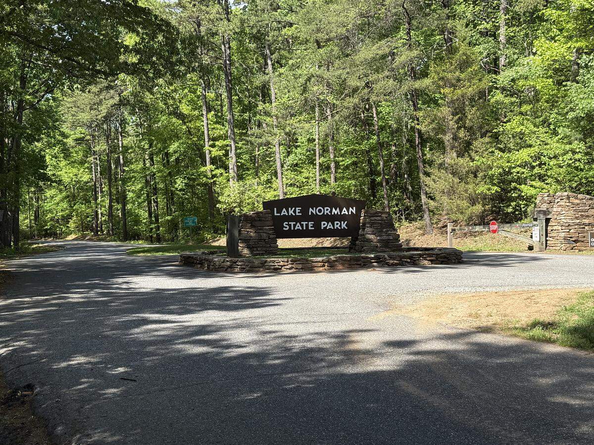 The rustic, stacked-stone entrance sign for Lake Norman State Park stands at a fork in an asphalt road, surrounded by a dense green forest on a sunny day.
