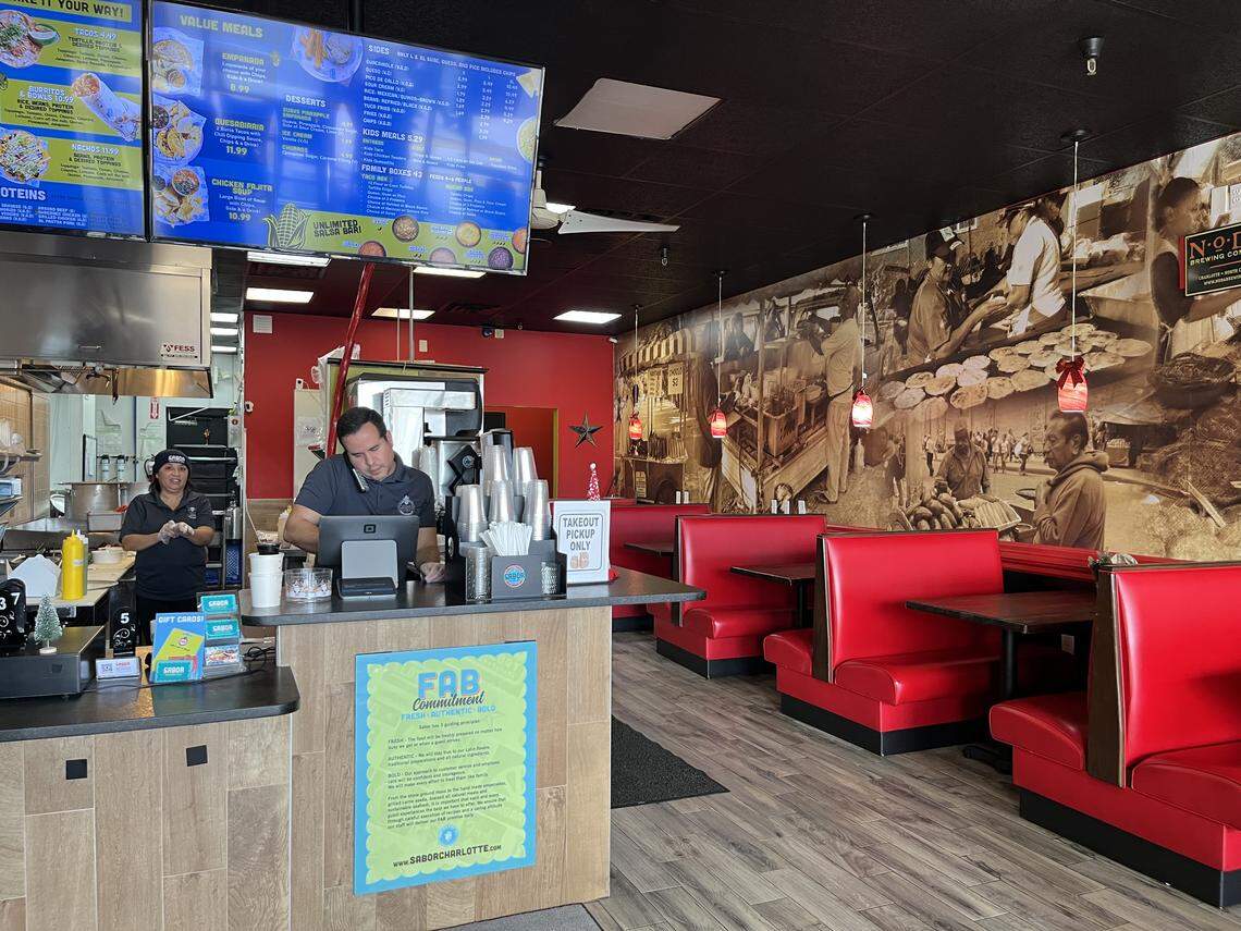 A wide, eye-level shot of a restaurant interior. Two employees are working behind the order counter under bright digital menu boards. The dining area features red vinyl booths and dark tables along a wall with a large, sepia-toned mural.