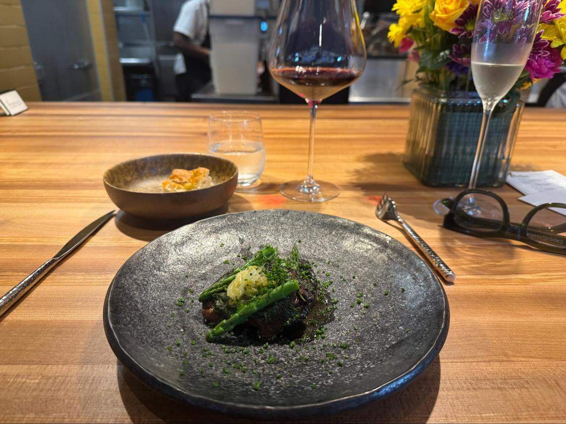 A wide shot of a chef’s counter featuring the seared protein dish from image 2. The wooden counter is also set with a glass of red wine, a small bowl of bread, a vase of yellow and purple flowers, and a pair of glasses.