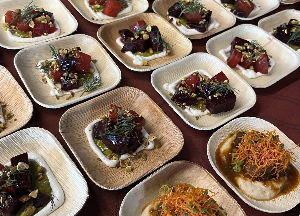 An overhead, slightly angled shot of several small, square bamboo plates arranged in rows on a dark red surface, likely at a catered event. The image features Beet Salad plates with cubes of deep red roasted beets resting on a white creamy base (possibly yogurt or labneh) and a vibrant green sauce (likely pesto or chimichurri). Each is garnished with crushed pistachios and a delicate sprig of fresh dill. Also shown are Braised Meat plates consisting of a mound of tender braised meat over a white purée, topped with a bright orange nest of crispy shredded vegetables (possibly sweet potatoes or carrots) and sliced chives.