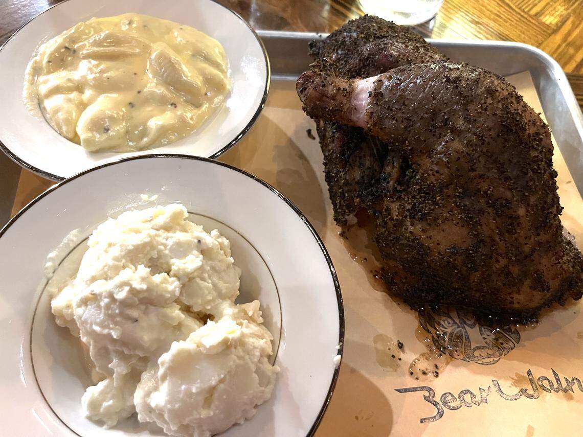 A high-angle shot of a meal on a paper-lined tray. The main course is a large piece of smoked chicken with a thick, coarse black pepper rub. It is accompanied by two white bowls containing side dishes of creamy potato salad and a cheesy mac and cheese.