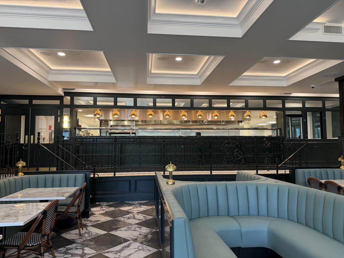 An interior view of a restaurant showing curved teal booths, marble-topped tables, and a view of the open kitchen pass with copper heat lamps.