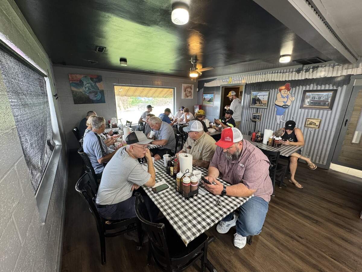 A high-angle, wide shot of the small, crowded dining room inside Boss Hog BBQ. Several patrons sit at long tables covered in black-and-white checkered tablecloths, many of them looking at their phones or eating. The room features dark wood flooring, gray concrete block walls, a corrugated metal accent wall with cartoon pig decor, and a large window looking out to the parking lot.