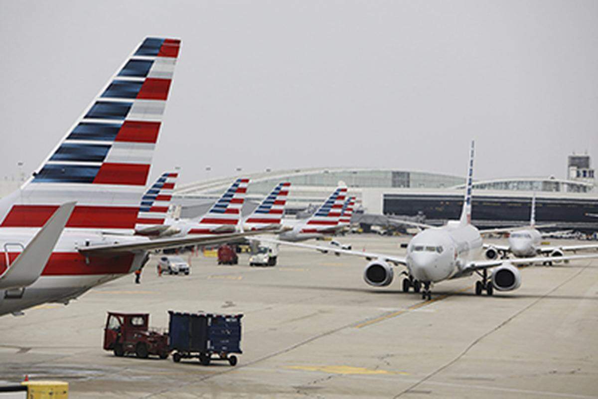 A wide view of an airport ramp shows the vertical stabilizers (tails) of several American Airlines jets lined up at gates on the left. On the right, a single American Airlines jet is taxiing in the foreground, with airport buildings and a control tower visible in the distance.