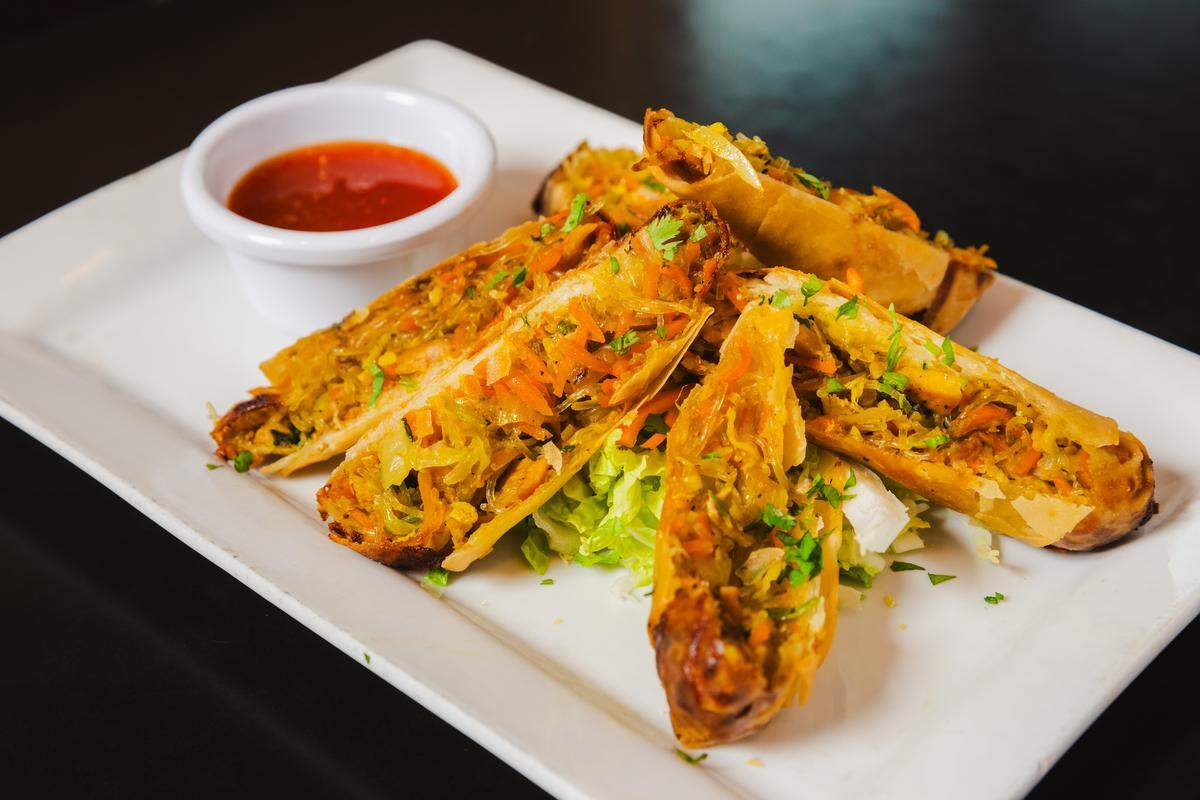 A plate of four crispy, fried smoked curry rolls sliced on a sharp diagonal to reveal a vibrant filling of shredded carrots, cabbage, and herbs. They are garnished with fresh cilantro and served on a bed of shredded lettuce with a small white ramekin of sweet chili sauce on a rectangular white plate.
