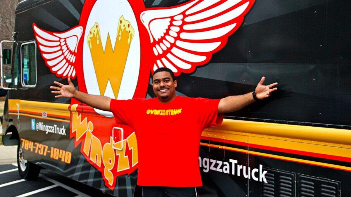 A smiling man stands with his arms outstretched in front of the black Wingzza food truck. He is wearing a red t-shirt with the truck’s logo, which features a large ‘W’ with red and white wings.