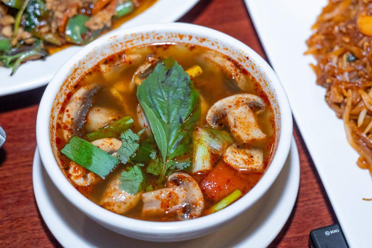 A detailed close-up of a bowl of Tom Yum soup. The clear, spicy broth is filled with sliced mushrooms, tomatoes, and green onions, topped with a large, vibrant green basil leaf and fresh cilantro.
