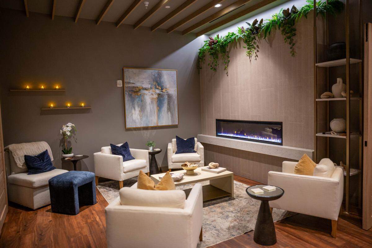 A wide-angle shot of a cozy, modern spa lounge featuring white upholstered armchairs arranged around a central coffee table on a patterned rug. A long, linear fireplace with blue flames is built into a wood-paneled wall, accented by hanging green plants above it. The room is decorated with abstract art, small side tables, and shelves holding vases and candles.