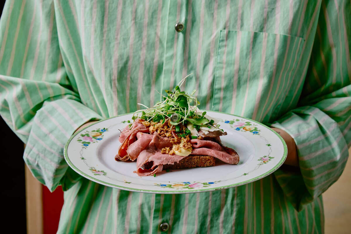 A person in a green striped shirt holding a floral vintage plate with a roast beef open-faced sandwich topped with microgreens.
