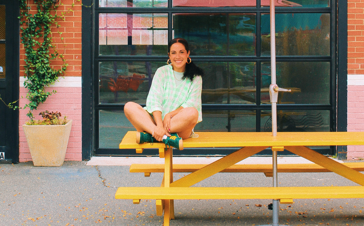 A woman with a dark ponytail sits cross-legged on a bright yellow picnic table, smiling directly at the camera. She is wearing a light green and white checkered shirt, shorts, and green shoes, and is in front of a brick building with large glass windows.