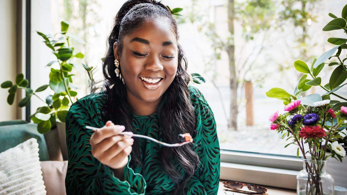 A smiling young woman is captured in a candid moment as she enjoys her food, surrounded by a cozy café atmosphere with lush greenery and vibrant flowers.