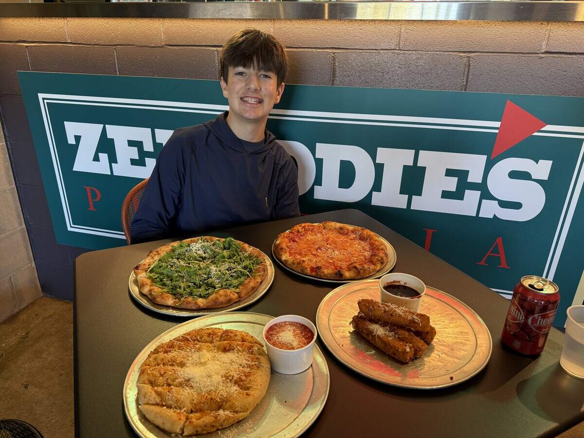 A smiling young person wearing a dark blue hoodie sits at a dark restaurant table holding a large spread of food. In front of them are four round metal trays displaying an arugula-topped pizza, a classic cheese pizza, a round cheesy garlic bread, and four thick breaded mozzarella sticks. There are also two small white cups filled with dipping sauce, a clear plastic cup, and a red can of Cheerwine cherry soda. Behind the person is a grey cinder block wall featuring a large green sign with partial white lettering for “Zepeddies Pizzeria.”