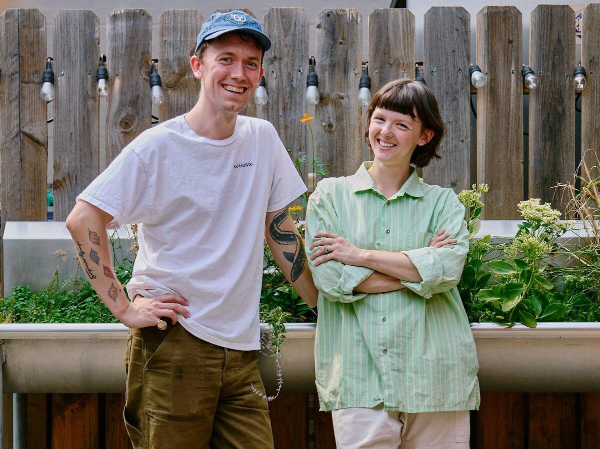 A person in a blue cap and a person in a green striped shirt smiling and standing together outside in front of a wooden fence.